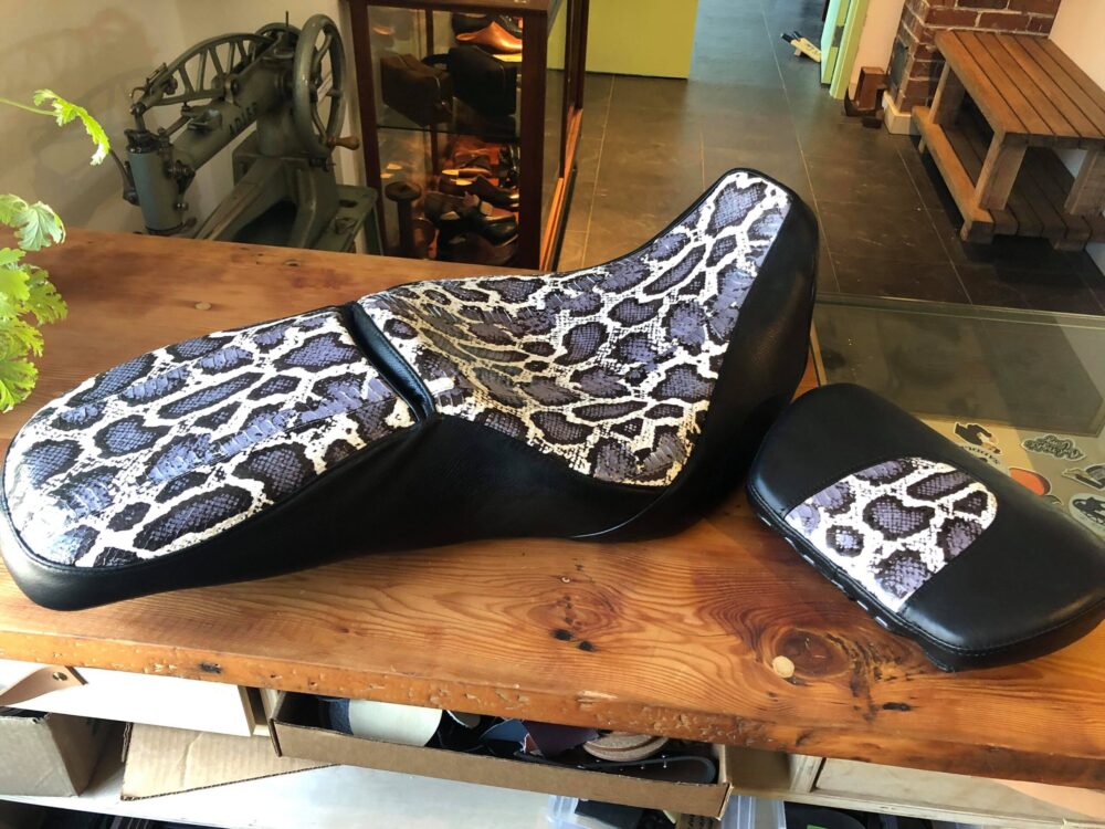 A custom upholstered motorcycle seat and headrest with blue-grey snakeskin and black leather sit on a wooden table top.
