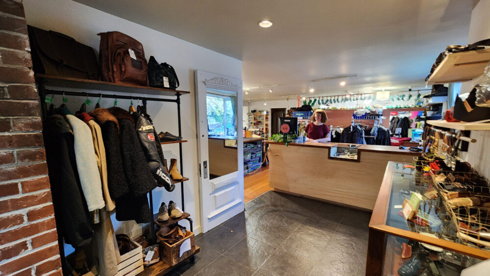 The entrance and retail area of the shop. There are racks with leather garments for sale and a mirror on the left, to the right there is a cabinet and shelves of handmade leather good for sale. Straight ahead is Tess standing behind the waist high wooden service counter.