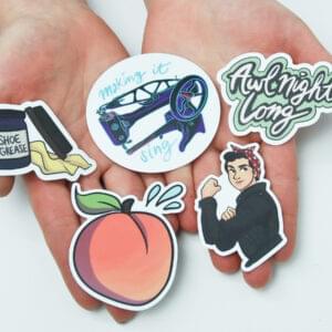 A pair of hands holding 5 illustrated stickers. From left to right, top to bottom, a jar of shoe grease with a cloth, a long arm sewing machine and "making it sing", "awl night long" on a green background, a peach with 3 dew drops coming off the top, a vintage "We Can Do It!" inspired figure with dark hair in a red bandana and black coveralls.