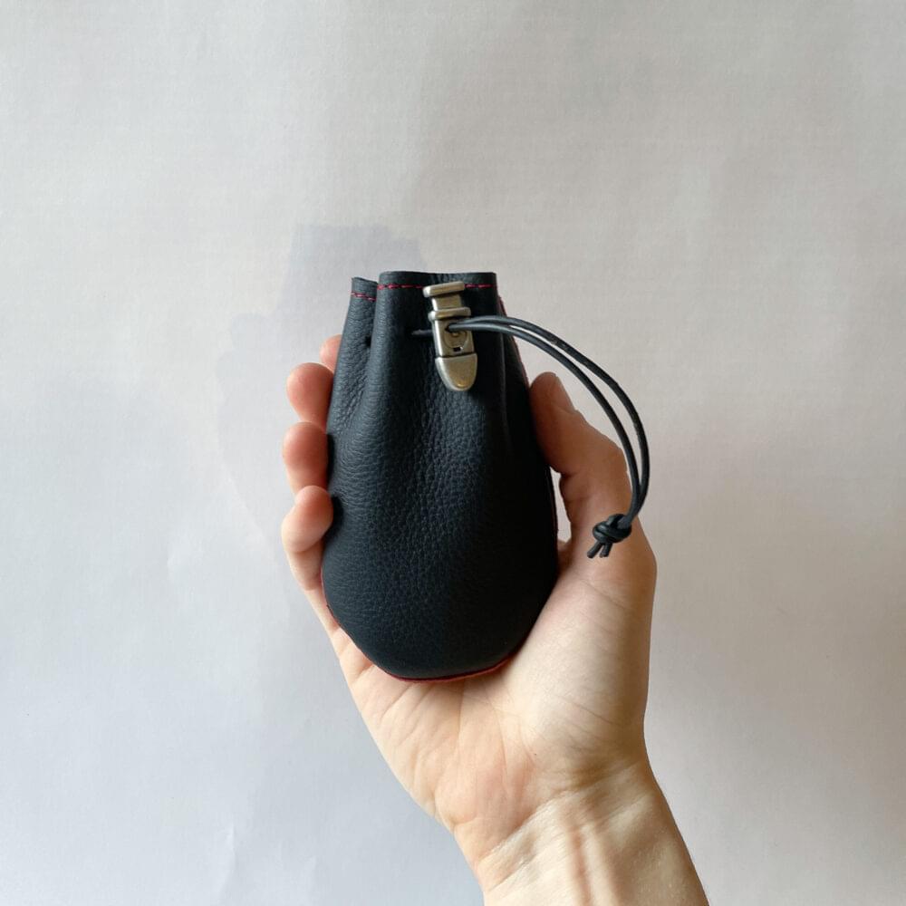 A small black leather drawstring pouch with red stitching, red piping, a black drawstring and an antique nickel cord lock, held by a hand with a plain light background.