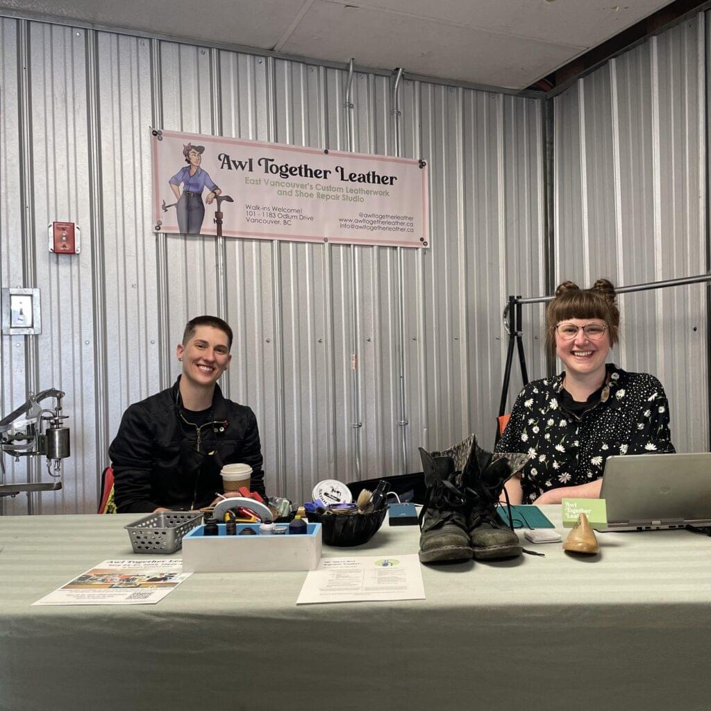 Ariss and Tess smiling for a photo taken sitting in Raven Recycling. There is a pink Awl Together Leather banner hanging on the wall behind them and sitting on a table in front of them are various items including tools for repairs, a pair of boots, a shoe last, and a laptop.