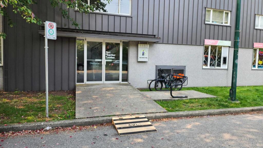 The entrance to the building at 1183 Odlum Drive from the street with a wooden ramp at the curb at the front glass double doors.